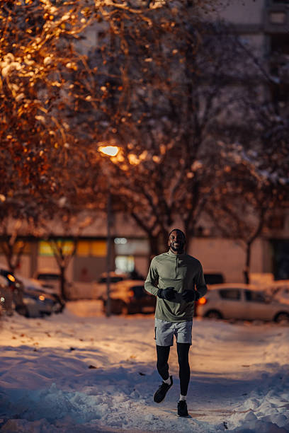 Young man jogging through snowy city streets at night, smiling and bundled in warm sportswear while staying fit and determined