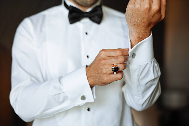 A young Caucasian man adjusts his cufflinks while wearing a formal black vest and white shirt. He stands indoors near a plant, preparing for an event.