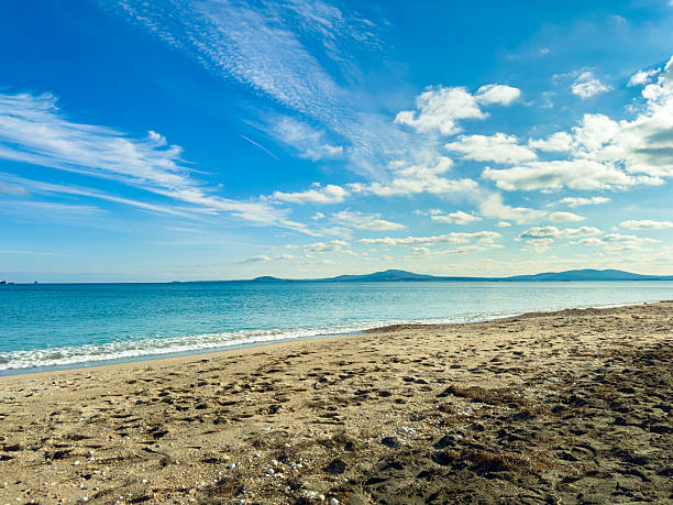 A beach scene shows soft sand, gentle waves, and a bright sky. The sun shines down, creating a tranquil setting with clear water and distant hills in the background.