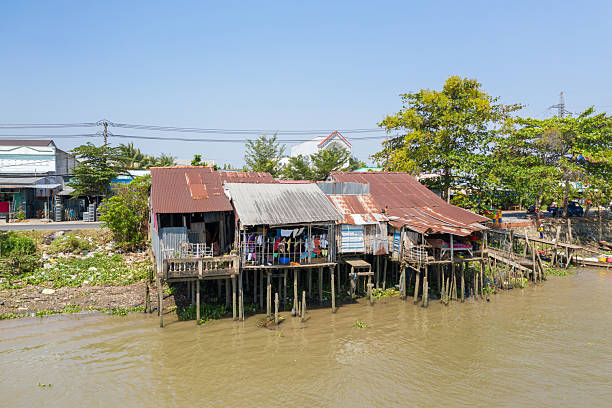Corrugated metal stilt houses with rusted roofs line the muddy riverbank at Cai Rang in Can Tho, Mekong Delta, Vietnam. Laundry hangs from balconies while lush trees and a clear blue sky frame the riverside settlement in bright daylight.
