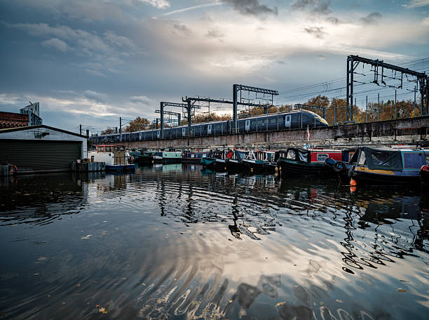 Wide urban scene showing boats docked along a canal with a train moving above on railway tracks. Reflective water, cloudy sky, and industrial surroundings illustrate contemporary city infrastructure and transport systems.