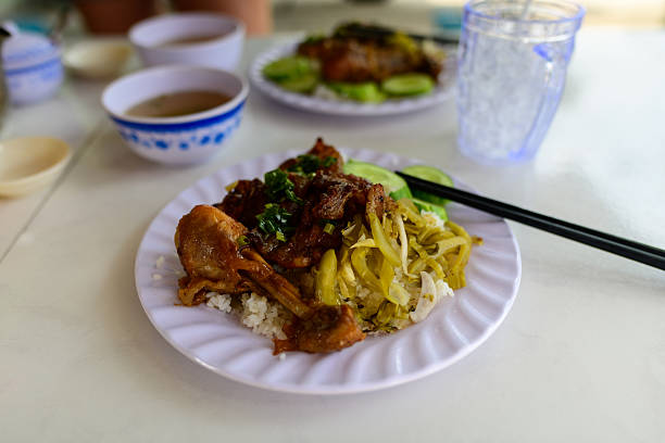 A close-up of a traditional Vietnamese rice dish featuring braised meat, pickled mustard greens, and sliced cucumber on a white plate. The meal is served with chopsticks and accompanied by bowls of soup and iced water on a simple table.
