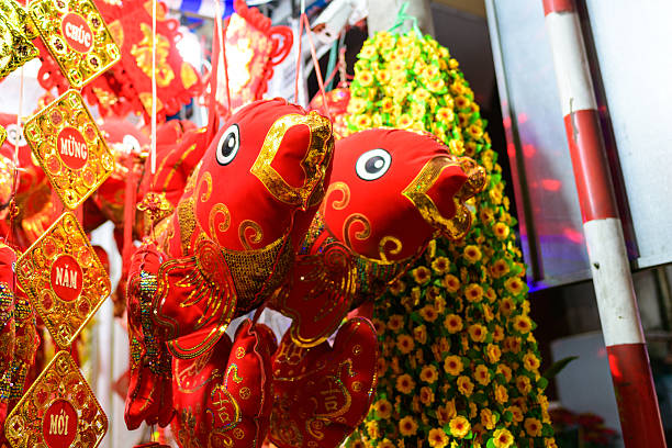 Brightly colored plush fish ornaments with gold embroidery hang alongside golden Tet banners and artificial yellow flowers at a bustling night market in Can Tho, Mekong Delta, southern Vietnam. The scene is illuminated by vivid market lights, highlighting the festive atmosphere and rich textures of the decorations.