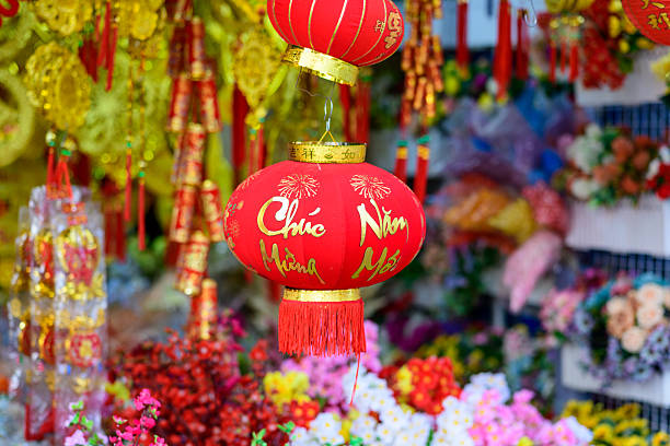 A vibrant red lantern decorated with gold calligraphy and fireworks motifs hangs among festive decorations at a market in Ben Tre, Mekong Delta, southern Vietnam. The scene features rich textures, bright colors, and a lively display of flowers and traditional ornaments for Lunar New Year.
