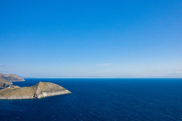 Minimal aerial composition featuring a sunlit, rocky peninsula extending into the expansive deep blue Mediterranean at Cap Tenare, Greece. The scene highlights open water, dry terrain, and a clear, cloudless sky for a sense of freedom and isolation.