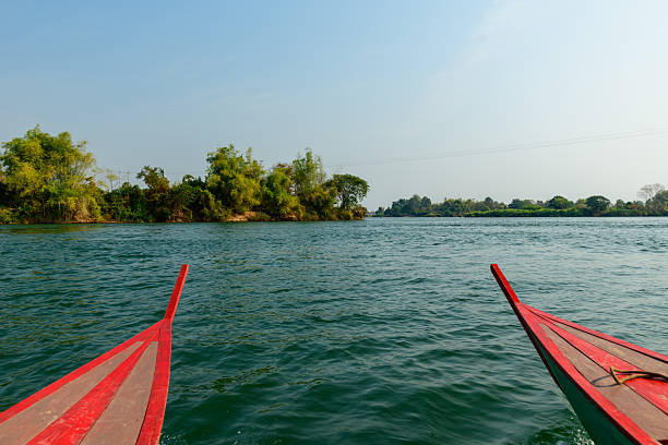 Two wooden boat prows with red trim point toward lush green islets on the Mekong River in the 4000 Islands region of southern Laos. Clear daylight illuminates the rippling water and the natural riverside vegetation under a wide blue sky.