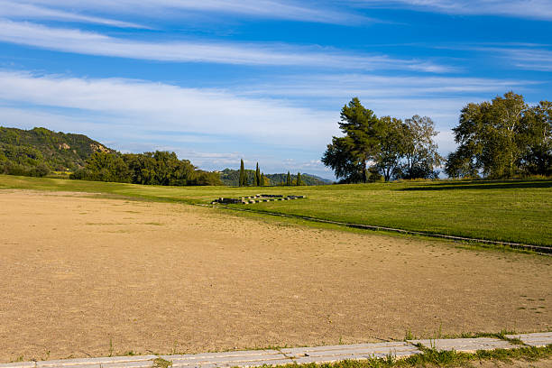 Open sandy track of the historic Olympic stadium in Olympia, Greece, bordered by grassy embankments and framed by distant hills and trees. Bright sunlight and a vivid blue sky highlight the expansive archaeological site and natural surroundings.