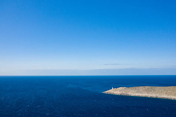 Wide aerial view of a solitary lighthouse on a rocky cape extending into deep blue Mediterranean waters at Cap Tenare, Greece. The scene features expansive open sea, clear sky, and a sense of isolation and tranquility.