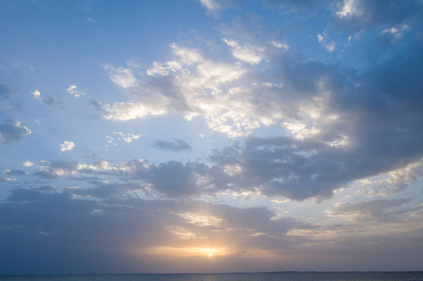 Gentle sun setting beneath scattered clouds casts pastel hues across the calm Mediterranean waters near Oristano, Sardinia. The peaceful sky and subtle light create an airy, serene atmosphere.