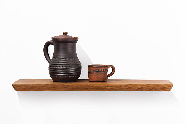 Clay jug with a handle and a mug standing on an oak wooden shelf attached to a white wall, front view.