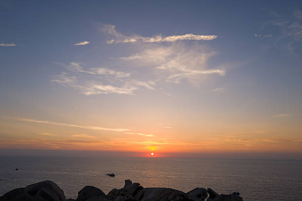 Gentle hues of orange and pink spread across the sky as the sun sets over the calm Mediterranean Sea, with smooth granite rocks silhouetted in the foreground at Capo Testa, Sardinia.