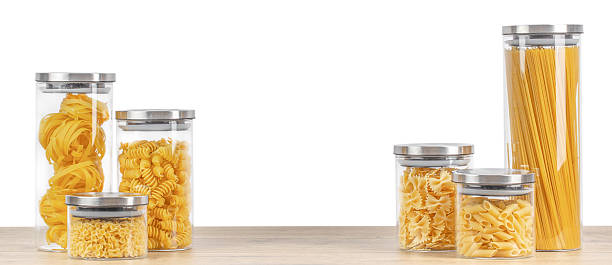 Collection of different types of dry pasta in clear glass storage jars with metal lids arranged on wooden surface, isolated, ideal for kitchen organization, food storage and grocery concepts