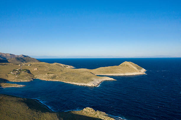 Aerial capture of a rugged peninsula jutting into the vivid blue Mediterranean at Cap Tenare, Greece. The windswept landscape meets open water beneath a clear sky, evoking a sense of wild seclusion.