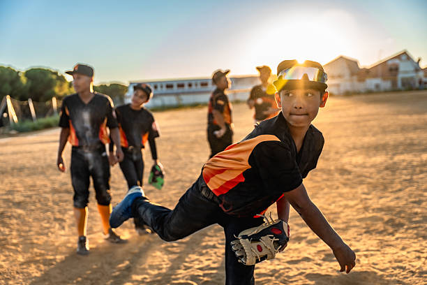 Young boy in baseball uniform and goggles pitches on a dusty field at sunset while teammates practice in the warm golden-hour light, focused and determined in play