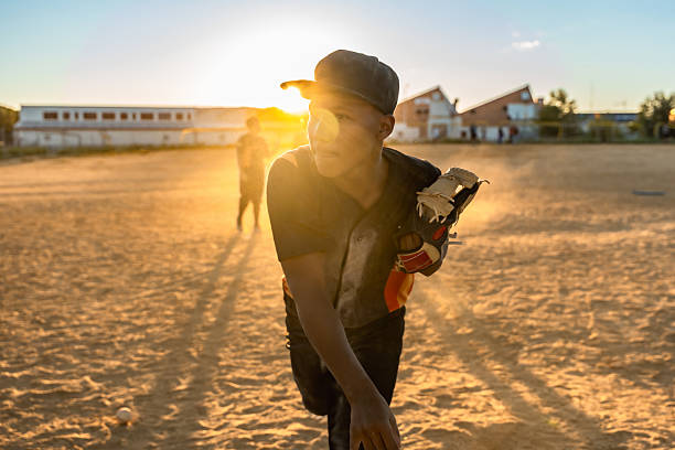 Young boy in baseball uniform wearing a glove, pitching during practice on a dusty field with a striking sunset creating a strong backlight and golden hour glow