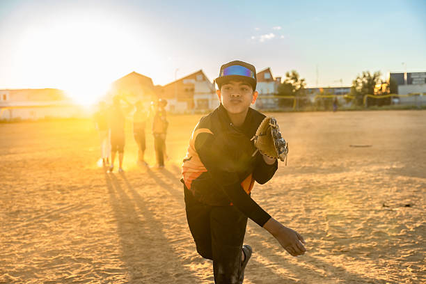 Young baseball player pitching on a dusty field during sunset, wearing a glove and uniform, with other blurred players and buildings in the warm background