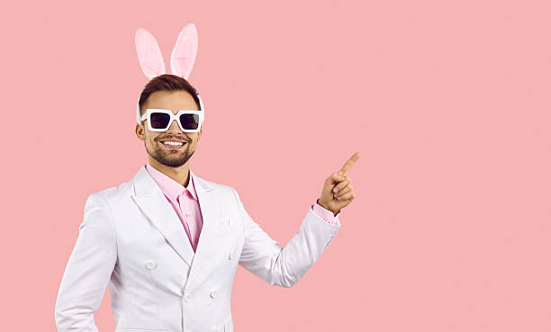 Smiling young man in suit and bunny ears isolated in pink background point at empty blank copy space aside. Happy male in costumer demonstrate deal or sale, recommend service. Ad concept.