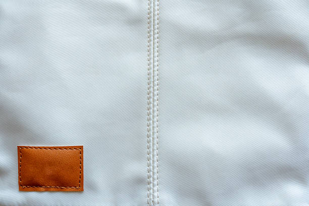 Close up of a light colored synthetic fabric featuring prominent vertical double stitching and a small rectangular blank brown leather patch in the lower left corner