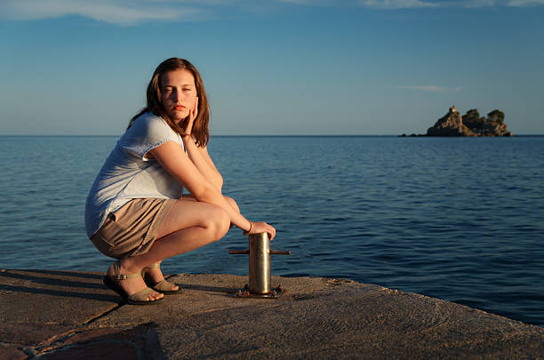Young woman in a crouched pose on a stone pier in Petrovac, Montenegro. Serious expression, hand on face, medium shot, sunset light, mediterranean travel.