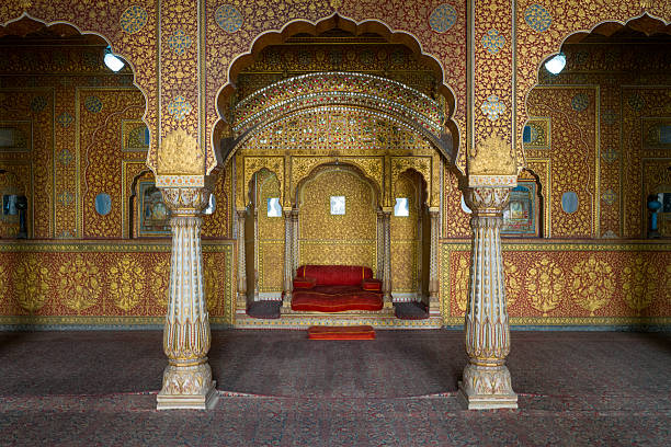 Lavishly decorated reception hall with intricate gold patterns, ornate arches, and a red velvet seat at Junagarh Fort in Bikaner, Rajasthan. Rich textures, detailed craftsmanship, and ambient natural light create a regal, historic atmosphere.