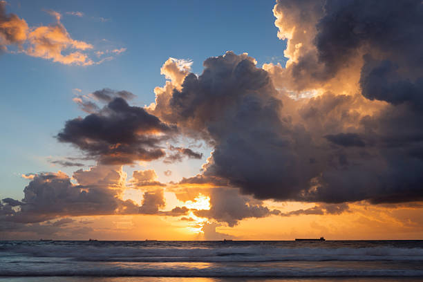 Huge vibrant moody clouds fill with light and colour from a golden ocean sunrise. Captured from the shores of Wollongong on the Illawarra East Coast of NSW, featuring silhouettes of cargo ships on the horizon.