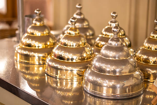shiny polished service cloches on counter, brass and silver domes reflecting warm ambient lighting, stacked in orderly row, upscale restaurant preparation,