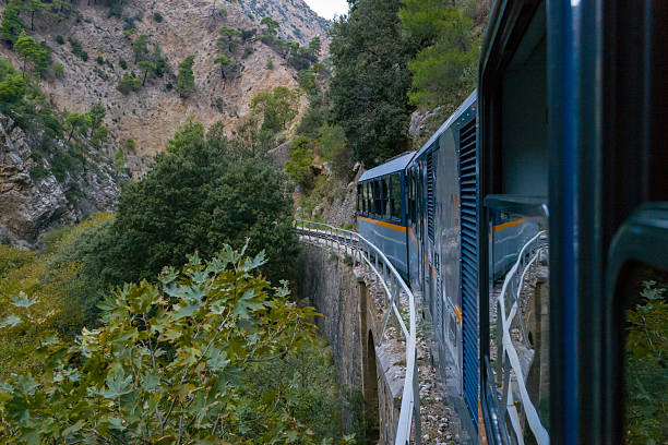 A blue narrow-gauge train travels along a curved stone bridge through the lush greenery and rugged cliffs of Vouraikos Gorge near Kalavrita, Greece. The scene features dense foliage, rocky slopes, and the dramatic landscape of the Peloponnese mountains.