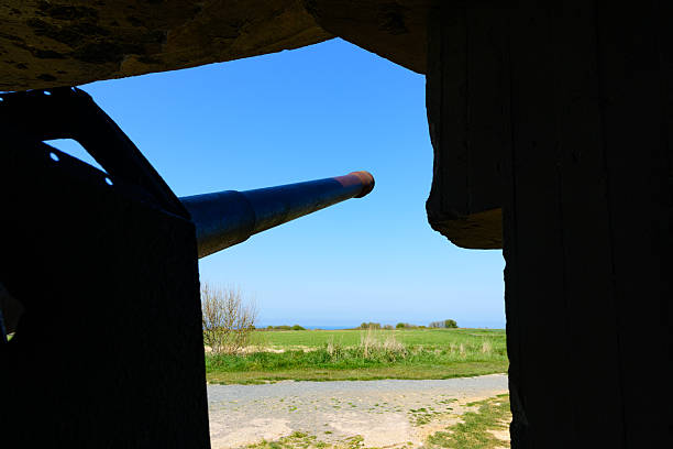 Looking out from inside a concrete World War II bunker at Longues-sur-Mer, Normandy, a large artillery gun barrel points toward open fields and a distant blue sky. Harsh shadows contrast with the bright landscape outside.