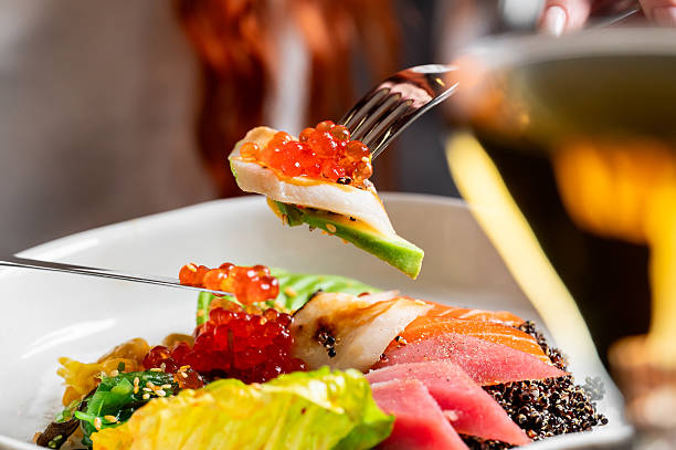 Close-up of a person using a fork to eat a fresh poke bowl with tuna, salmon, avocado, seaweed, and red roe over black rice