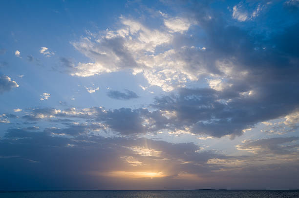 Expansive view of the Mediterranean near Oristano, Sardinia, with dramatic clouds illuminated by soft evening light above tranquil water. The serene seascape features gentle rays breaking through the sky at sunset.