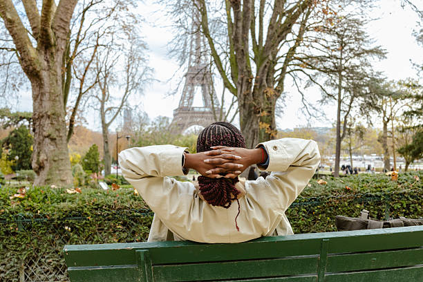 A relaxed young woman sits on a green park bench in Paris, hands behind her head, enjoying a sunny day with the Eiffel Tower visible in the distance.