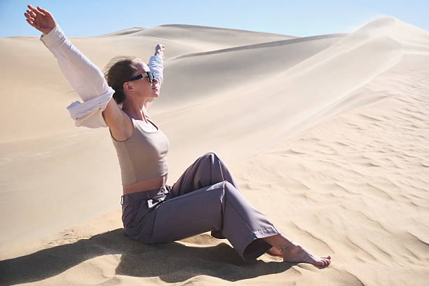 Young woman sitting on soft sand dunes with raised arms, enjoying open desert landscape under bright sunlight. Freedom, joy, mindfulness, travel. Sand dunes in Sahara desert, Siwa oasis, Egypt