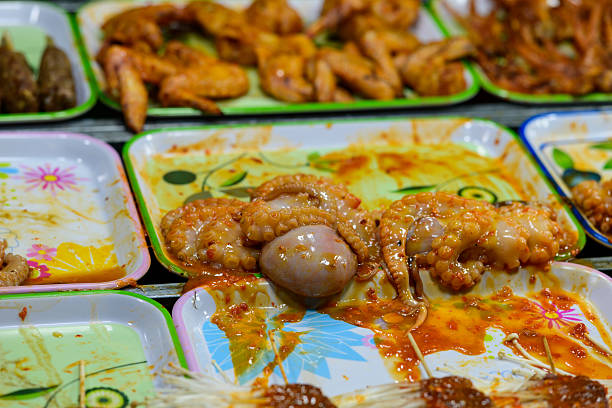 Close-up of raw octopus tentacles coated in spicy marinade, displayed on colorful trays alongside other street food at the bustling night market in Can Tho, Mekong Delta, southern Vietnam. The scene features vibrant sauces, varied textures, and a lively food stall atmosphere.