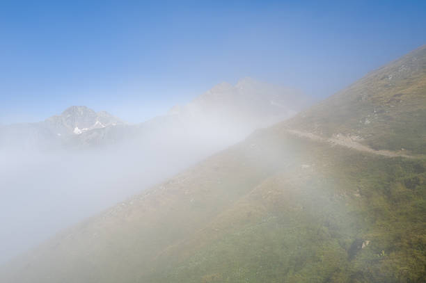 A narrow hiking path curves along a grassy mountain slope as thick fog drifts across the landscape, partially obscuring distant peaks beneath a clear blue sky.