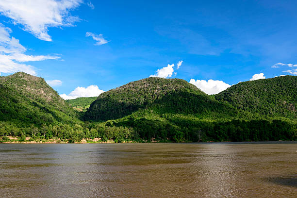 Bright sunlight falls across dense green hills rising above the wide Mekong river, with vivid blue sky and scattered white clouds overhead. The scene highlights lush tropical vegetation and the tranquil flow of water in Southeast Asia.
