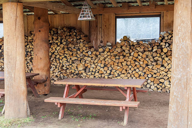 Outdoor wooden picnic table and benches under a shelter with a large wall of stacked firewood. Concept for rural tourism, cozy rest stops, and country lifestyle.