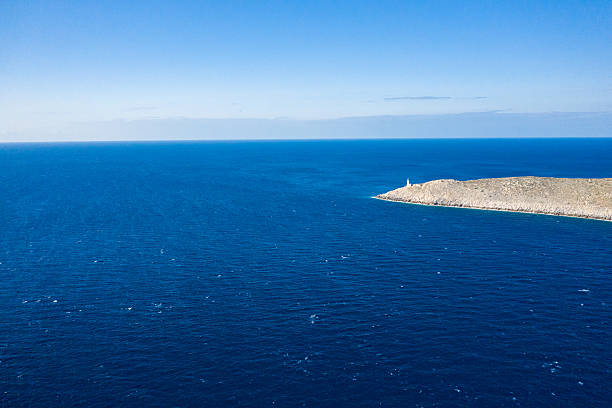 A solitary lighthouse stands at the tip of a barren, rocky peninsula surrounded by deep blue Mediterranean waters at Cap Tenare, Greece. The scene is sunlit and expansive, with vivid ocean hues and a clear sky evoking isolation and tranquility.