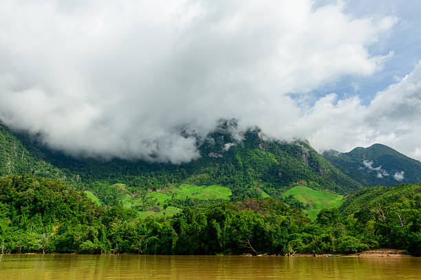 Thick clouds descend over lush, forested hills above the muddy Nam Ou River near Muang Ngoi in northern Laos. The vibrant landscape features dense jungle, rolling green slopes, and a dramatic sky, creating a serene natural scene.