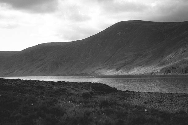 Black and white view of a loch surrounded by rolling hills under an overcast sky.