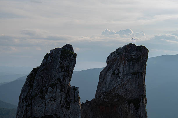 A dramatic mountain landscape featuring a cross on a high rocky pinnacle, with another peak nearby. The distant ridges and soft clouds create a tranquil, contemplative outdoor scene perfect for nature and spirituality themes.