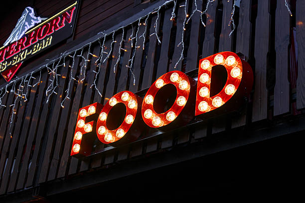 Looking up at a neon sign spelling “foods”, sits on a roof of a restaurant in the day with a puffy cloud against a blue sky.