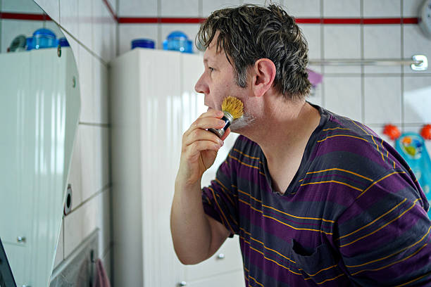 Young man shaving in a blue bathroom, carefully removing hair to reveal smooth skin. His daily grooming routine ensures healthy skin