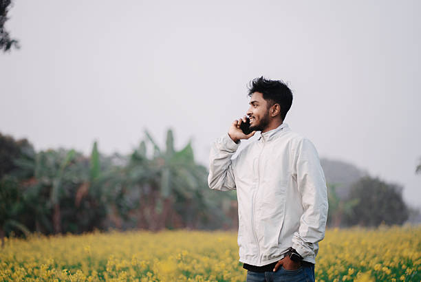 Young Indian man talking on a mobile phone while standing in a mustard flower field. Concept of communication, rural connectivity, mobile technology, and modern India.