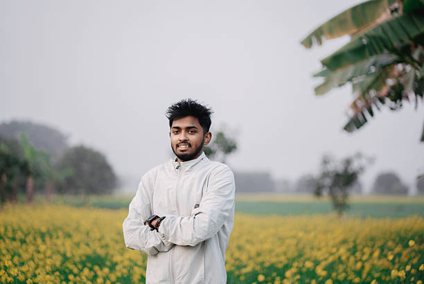 Confident young Indian man standing with crossed arms in a mustard flower field. Concept of rural lifestyle, confidence, youth, and modern India.