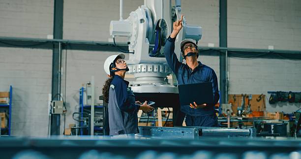 istock Two young engineers, one using laptop and the other handheld device, collaborate in factory to manage robotic arm. Robotic Industrial Factory. 2255686436