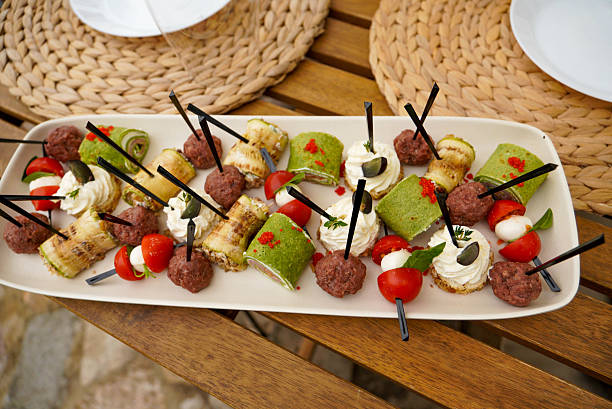 Elegant gourmet appetizer platter featuring assorted finger food made from local ingredients. Neatly arranged bite-sized snacks including meatballs, cheese cream canapés, cherry tomatoes, olives and green savory rolls, served on a white plate. Perfect for catering, events, restaurant menus and food presentation concepts.