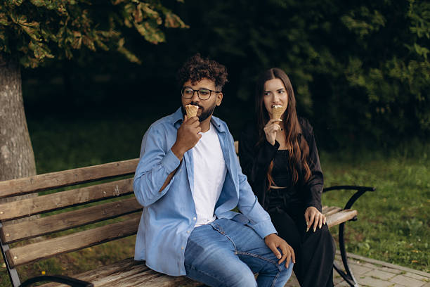 Happy interracial couple relaxing on a bench, eating ice cream cones outdoors, enjoying a sweet summer dessert together