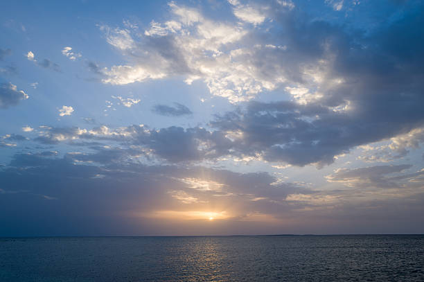 Expansive view of the Mediterranean Sea near Oristano, Sardinia, with dramatic clouds and warm sunlight breaking through at sunset. Soft reflections shimmer on calm water, creating a tranquil, spacious mood.
