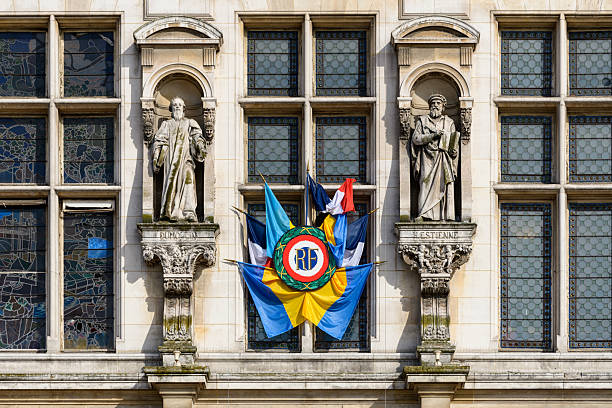 Two stone statues stand in niches above a vibrant cluster of civic flags on the ornate facade of Hotel de Ville in Paris, framed by stained glass and patterned windows in bright sunlight.