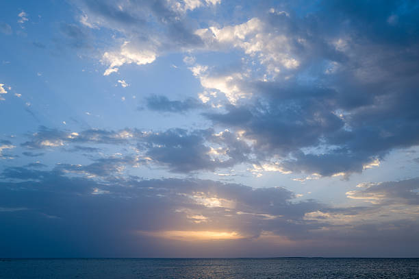 Soft pastel clouds drift across a serene sky as the sun sets over calm Mediterranean waters near Oristano, Sardinia. Gentle light and subtle gradients create a peaceful, airy seascape.